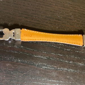 May include: Close-up of a silver-toned belt with a tan leather section. The leather is stitched with white thread and has the word "CHINA" stamped on it. The belt has decorative clover-shaped links.