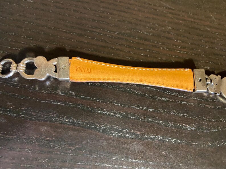 May include: Close-up of a silver-toned belt with a tan leather section. The leather is stitched with white thread and has the word "CHINA" stamped on it. The belt has decorative clover-shaped links.