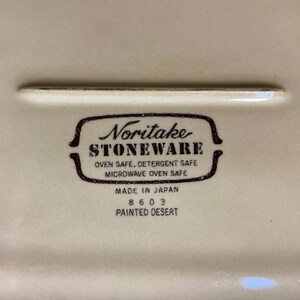 May include: Close-up of a cream-colored ceramic dish with a brown Noritake Stoneware logo. The logo includes text indicating the dish is oven, detergent, and microwave safe. The dish is marked "Made in Japan" and "Painted Desert".