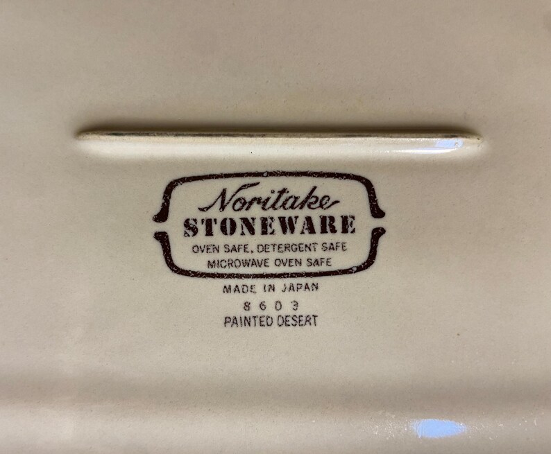 May include: Close-up of a cream-colored ceramic dish with a brown Noritake Stoneware logo. The logo includes text indicating the dish is oven, detergent, and microwave safe. The dish is marked "Made in Japan" and "Painted Desert".