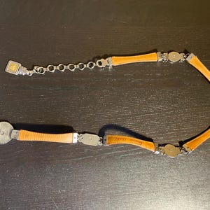 May include: A tan leather belt with silver-tone metal accents. The belt features rectangular and circular metal pieces connecting the leather segments. The buckle is a silver-tone metal with a decorative design.