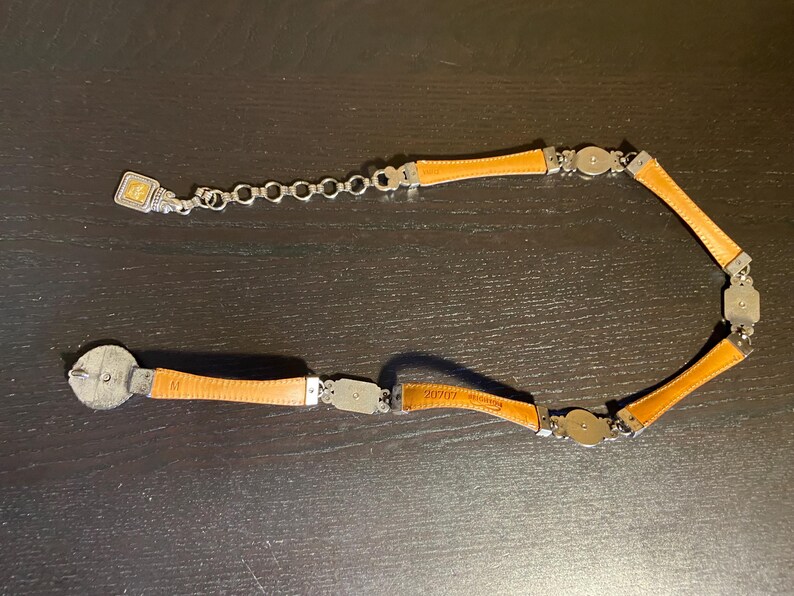 May include: A tan leather belt with silver-tone metal accents. The belt features rectangular and circular metal pieces connecting the leather segments. The buckle is a silver-tone metal with a decorative design.