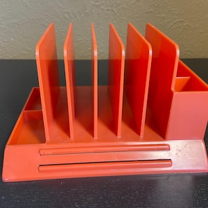 May include: A vibrant orange desk tidy with several vertical dividers, a small compartment, and a lower tray. The organiser is made of plastic and is designed to hold papers, pens, and other office supplies.