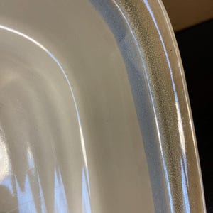 May include: Close-up of a white ceramic dish with a rounded edge. The rim features a speckled, textured band in shades of brown and blue. The dish has a glossy finish.