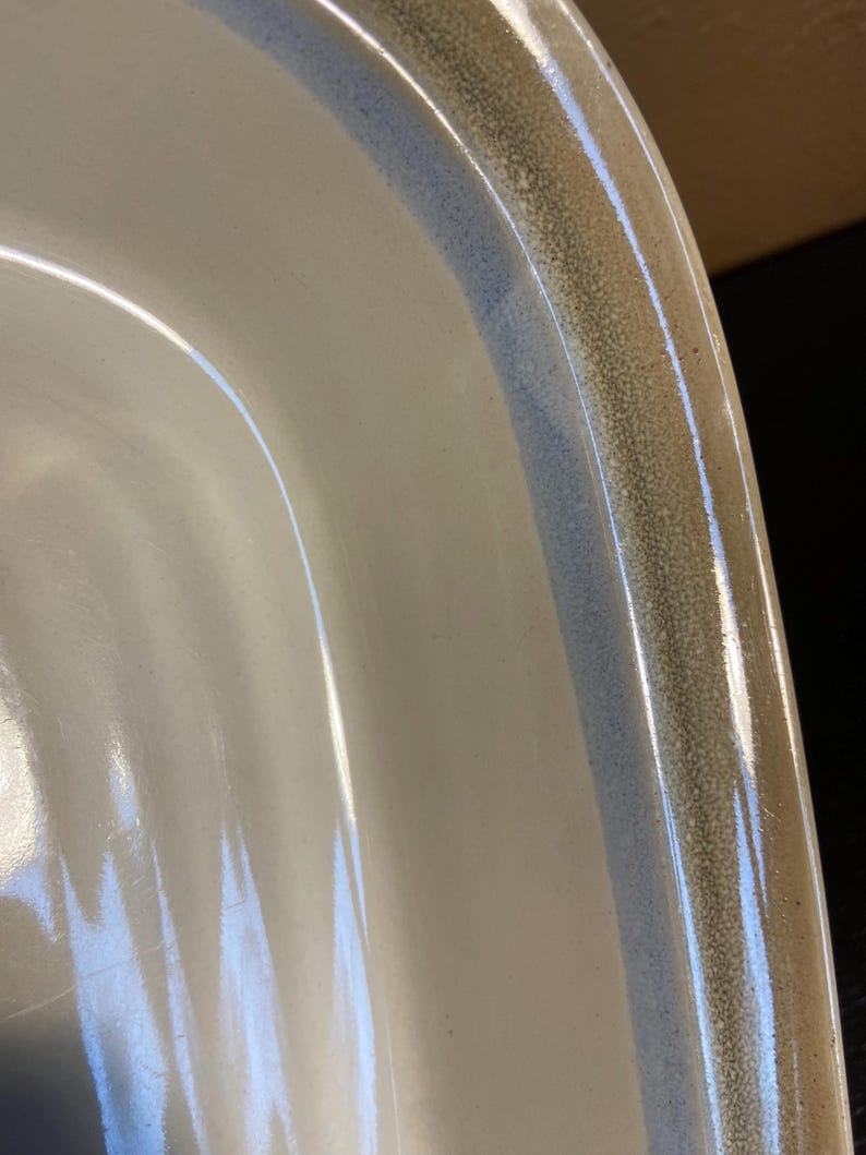 May include: Close-up of a white ceramic dish with a rounded edge. The rim features a speckled, textured band in shades of brown and blue. The dish has a glossy finish.