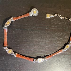 May include: A brown leather belt with silver and gold-toned metal accents. The belt features a series of rectangular and circular decorative elements. The belt is designed with a clasp closure and a chain for adjusting the size.