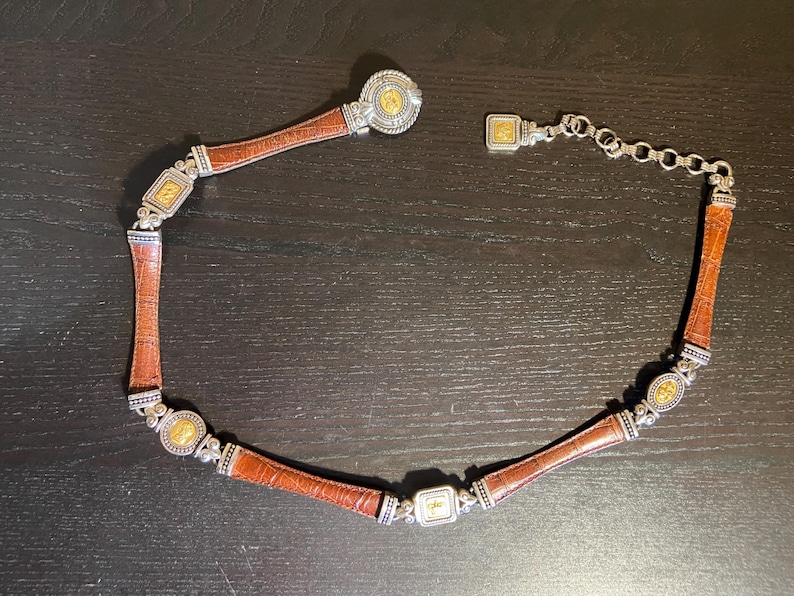 May include: A brown leather belt with silver and gold-toned metal accents. The belt features a series of rectangular and circular decorative elements. The belt is designed with a clasp closure and a chain for adjusting the size.