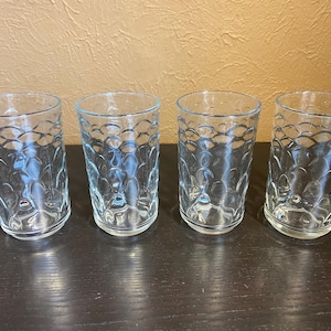 Puede incluir: Cuatro vasos transparentes con un patrón texturizado en forma de burbujas. Los vasos son cilíndricos con una parte superior ligeramente ensanchada. Están sobre una superficie oscura.