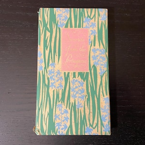 May include: A vintage book with a green cover featuring a floral pattern of blue flowers and gold accents. The center has a pink rectangle with the title "From the Porch". The book is closed and resting on a dark wooden surface.