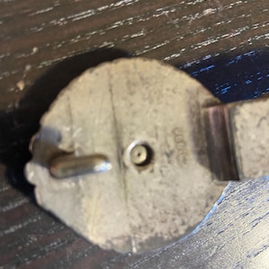 May include: Close-up of a silver-toned metal belt buckle. The buckle has a decorative, possibly floral, edge and a central pin for closure. The metal shows signs of wear, suggesting age or use. The buckle is set against a dark wooden surface.