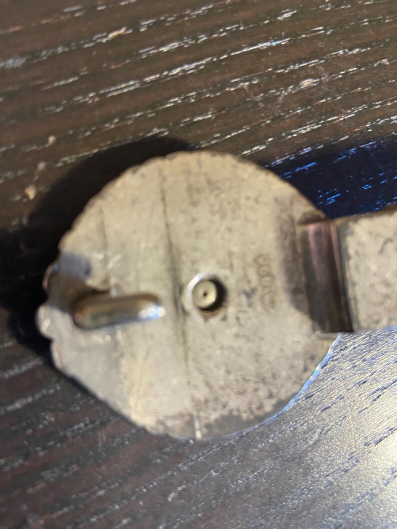 May include: Close-up of a silver-toned metal belt buckle. The buckle has a decorative, possibly floral, edge and a central pin for closure. The metal shows signs of wear, suggesting age or use. The buckle is set against a dark wooden surface.