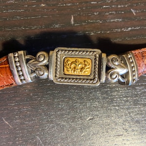 May include: Close-up of a brown leather belt with a silver and gold buckle. The buckle features a rectangular gold center with a fleur-de-lis design, framed by a silver border with rope detailing. The belt is a warm brown color.