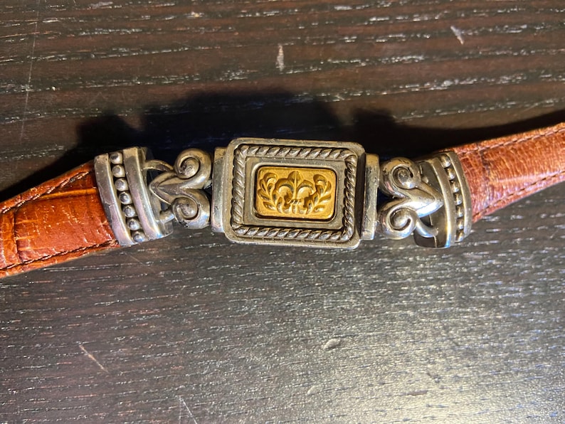 May include: Close-up of a brown leather belt with a silver and gold buckle. The buckle features a rectangular gold center with a fleur-de-lis design, framed by a silver border with rope detailing. The belt is a warm brown color.