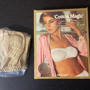Puede incluir: Una caja vintage y un sujetador de tubo. La caja presenta a una mujer con un sujetador de tubo blanco y una bata rosa. El texto en la caja dice "Cotton Magic" y describe el sujetador como elástico, con cierre trasero y 100% algodón. El sujetador es beige y está en una bolsa de plástico.