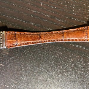 May include: A brown leather bracelet with a textured, crocodile-like pattern. The center leather section is flanked by silver-toned metal accents and decorative filigree. The bracelet is set against a dark wooden surface.