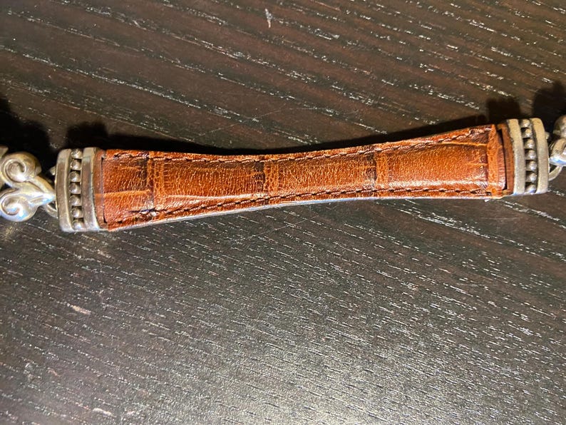 May include: A brown leather bracelet with a textured, crocodile-like pattern. The center leather section is flanked by silver-toned metal accents and decorative filigree. The bracelet is set against a dark wooden surface.