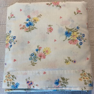 Puede incluir: Una sábana de algodón blanca con un patrón floral con flores rosas, azules y amarillas. Las flores están dispersas por la sábana en un patrón aleatorio.