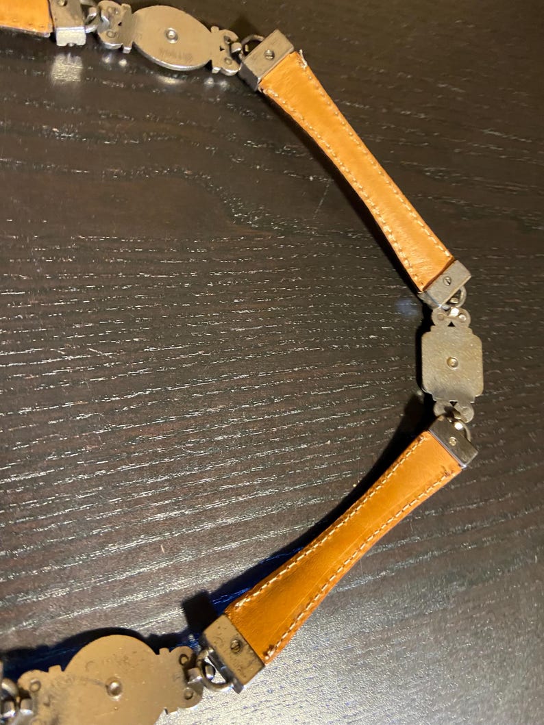 May include: A vintage belt featuring a combination of silver-toned metal and tan leather. The belt has a series of decorative metal links and rectangular leather sections. The leather is stitched with a contrasting thread. The belt is designed to be worn around the waist.