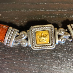 May include: A brown leather belt with a silver and gold buckle. The buckle features a gold crown design in a square frame. The belt has a textured, crocodile-like pattern.