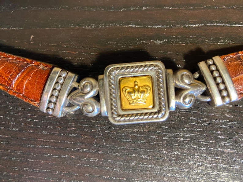 May include: A brown leather belt with a silver and gold buckle. The buckle features a gold crown design in a square frame. The belt has a textured, crocodile-like pattern.