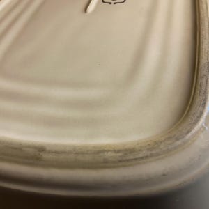 May include: Off-white ceramic serving dish with a slightly curved rectangular shape. The rim has a dark, aged appearance. The dish's surface has subtle, concentric ridges. The bottom of the dish is visible.