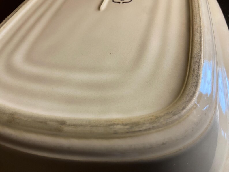 May include: Off-white ceramic serving dish with a slightly curved rectangular shape. The rim has a dark, aged appearance. The dish's surface has subtle, concentric ridges. The bottom of the dish is visible.