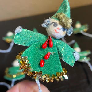 May include: A small elf ornament featuring a green felt body with gold tinsel trim, three red teardrop-shaped accents, and a pointed green hat. The figure has a round white head with painted facial features and fuzzy gray hair accents.
