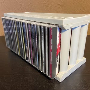 Könnte beinhalten: Ein weißer CD-Aufbewahrungskoffer aus Kunststoff, gefüllt mit CDs. Der Koffer hat eine rechteckige Form mit Deckel und wird von vier zylindrischen Säulen getragen. Die CDs sind sichtbar, ihre Buchrücken zeigen nach außen.