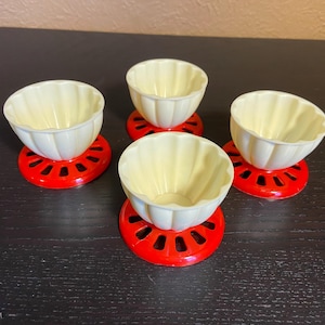 Könnte beinhalten: Vier cremefarbene, geriffelte Schalen stehen auf leuchtend roten, kreisförmigen Ständern. Die Schalen haben einen gewellten Rand und ein glattes Inneres. Die Ständer haben ein dekoratives, offenes Design. Die Artikel sind auf einer dunklen Oberfläche angeordnet.