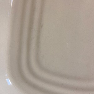 May include: Close-up of a light beige ceramic plate with a curved edge. The plate has a smooth surface with subtle variations in color and texture. The curved edge features three concentric ridges.