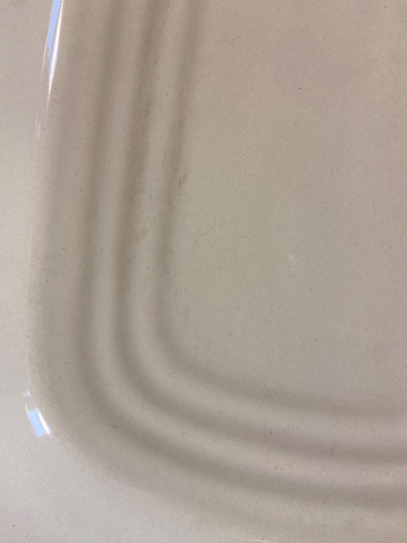 May include: Close-up of a light beige ceramic plate with a curved edge. The plate has a smooth surface with subtle variations in color and texture. The curved edge features three concentric ridges.