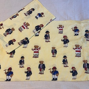 Pode incluir: Duas fronhas retangulares com um padrão de riscas verticais finas em amarelo claro e branco. O tecido é decorado com uma estampa repetitiva de ursos de peluche vestidos com várias roupas, incluindo blazers, camisolas e chapéus, em diferentes poses.