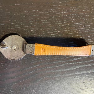 May include: A close-up of a belt with a silver buckle and a tan leather strap. The leather strap has the letter "M" embossed on it. The belt is on a dark wooden surface.