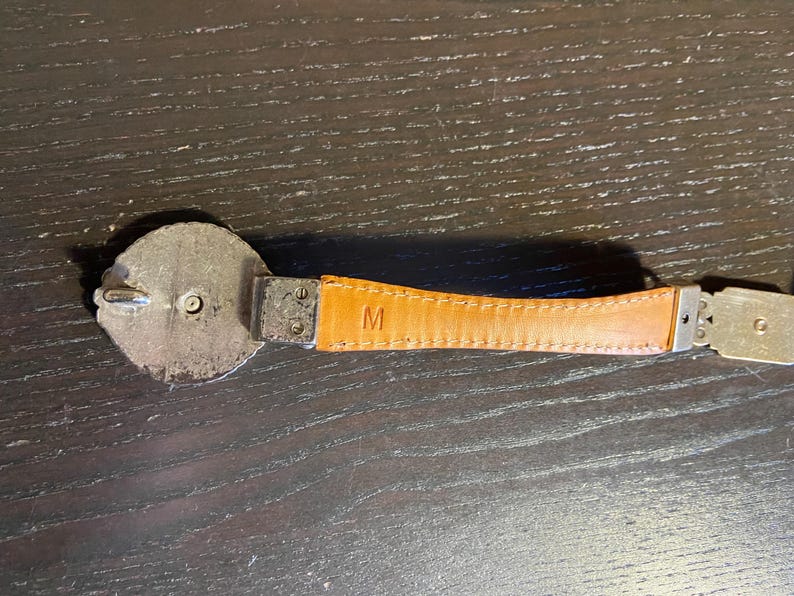 May include: A close-up of a belt with a silver buckle and a tan leather strap. The leather strap has the letter "M" embossed on it. The belt is on a dark wooden surface.