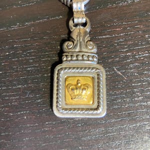 May include: Silver-toned pendant with a gold-colored square center featuring a crown design. The pendant has an ornate top and a braided border around the gold square. The pendant is attached to a silver chain.