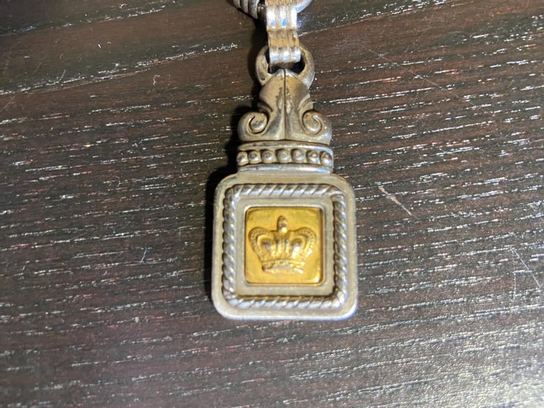 May include: Silver-toned pendant with a gold-colored square center featuring a crown design. The pendant has an ornate top and a braided border around the gold square. The pendant is attached to a silver chain.