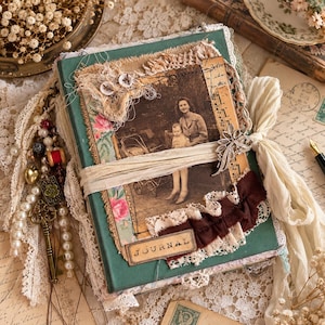 Puede incluir: Un diario verde con una fotografía antigua de una persona y un niño, decorado con encaje, retazos de tela y dos botones blancos. La cubierta incluye un adorno floral de metal, una cinta color crema y un colgante con cuentas de perlas.