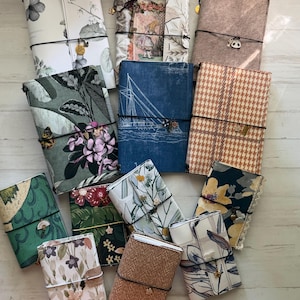 Peut inclure: Une collection de douze carnets rectangulaires recouverts de tissu, chacun fixé par un élastique noir et orné d'une petite breloque en métal. Les couvertures présentent divers motifs, notamment des imprimés floraux, pied-de-poule, nautiques et des textures unies.