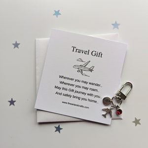 Può includere: Un biglietto bianco con testo nero che recita "Travel Gift" e una piccola illustrazione di un aereo. Il biglietto è piegato a metà e un portachiavi argentato con un piccolo ciondolo a forma di aereo e un strass rosso è attaccato al biglietto.