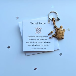 May include: A small wooden turtle keychain with a beaded cord and a silver charm. The keychain is accompanied by a card that reads "Travel Turtle" and a poem about safe travels.