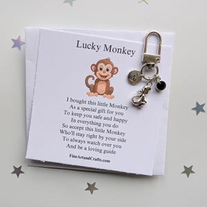 Könnte beinhalten: Eine weiße Karte mit dem Text "Lucky Monkey" und einem Gedicht über einen Affen-Schlüsselanhänger. Der Schlüsselanhänger besteht aus einem silbernen Affen-Anhänger, einem silbernen Buchstaben-Anhänger und einem schwarzen Perlen-Anhänger. Die Karte liegt auf einem weißen Hintergrund mit silbernen Sternen.