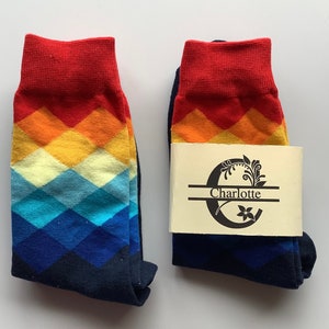 May include: A pair of colourful socks with a chevron pattern in shades of red, orange, yellow, blue, and navy. The socks have a personalised tag with the name "Charlotte" on it.