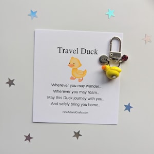 May include: A yellow rubber duck keyring with a silver charm and a red crystal charm. The keyring is attached to a white card with the text "Travel Duck" and a poem about safe travels.