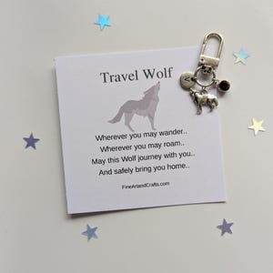 May include: A silver-toned metal keyring featuring a wolf charm, a small circular disc engraved with the letter V, and a dark red bead. It is displayed next to a white card printed with the text Travel Wolf and a poem about wandering.