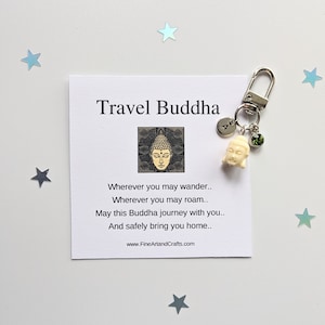 Op de afbeelding: Een witte reis-Boeddha sleutelhanger met een groene edelsteen charme en een zilveren letter charme. De sleutelhanger is bevestigd aan een kaart met de tekst "Travel Buddha" en een gedicht over veilig reizen.