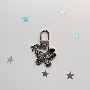 May include: Silver key chain with a butterfly charm, a small crystal, and the letter "R" hanging from it. The keychain is on a white background with scattered iridescent stars.