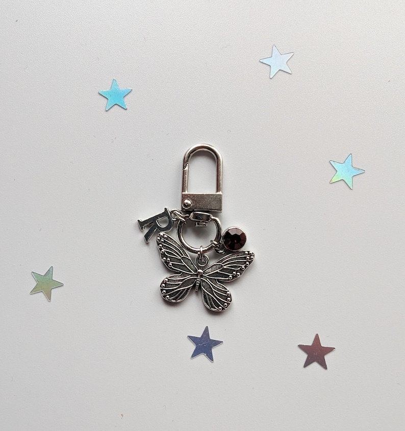 May include: Silver key chain with a butterfly charm, a small crystal, and the letter "R" hanging from it. The keychain is on a white background with scattered iridescent stars.