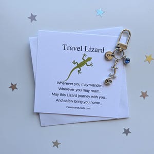 Könnte beinhalten: Ein silberfarbener Schlüsselanhänger mit einem Eidechsen-Anhänger und einem blauen Edelstein, befestigt an einer Karte mit der Aufschrift "Travel Lizard". Die Karte zeigt eine grüne Eidechsen-Illustration und ein Gedicht. Der Hintergrund ist hellblau mit sternförmigem Konfetti.