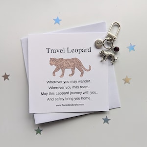 Puede incluir: Un llavero plateado con un dije de leopardo y una pequeña gema roja, junto a una tarjeta con el texto "Travel Leopard" y una ilustración de leopardo. La tarjeta incluye un poema. Estrellas decorativas están dispersas.