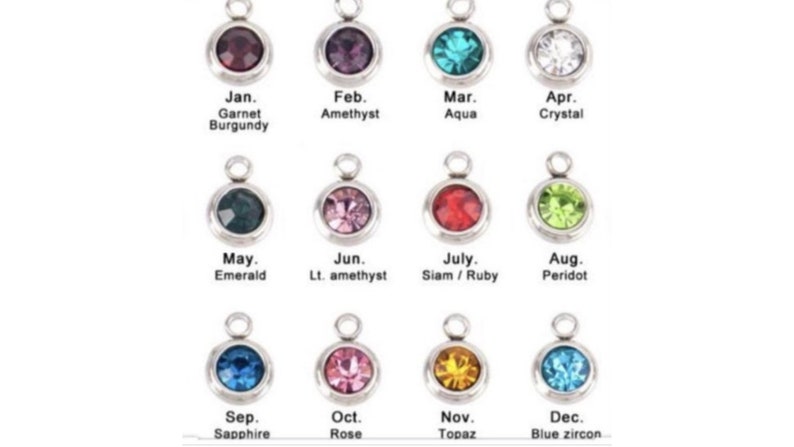 May include: Twelve silver charms with different colored gemstones, each representing a month of the year. The charms are labeled with the month and the corresponding birthstone.